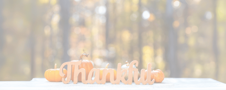 Blurred image of fall trees in rural America with thankful written in wood with a scroll saw and pumpkins of varying Colors and shapes behind it